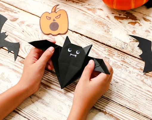 Child crafting origami bat with Halloween decorations on wooden table.