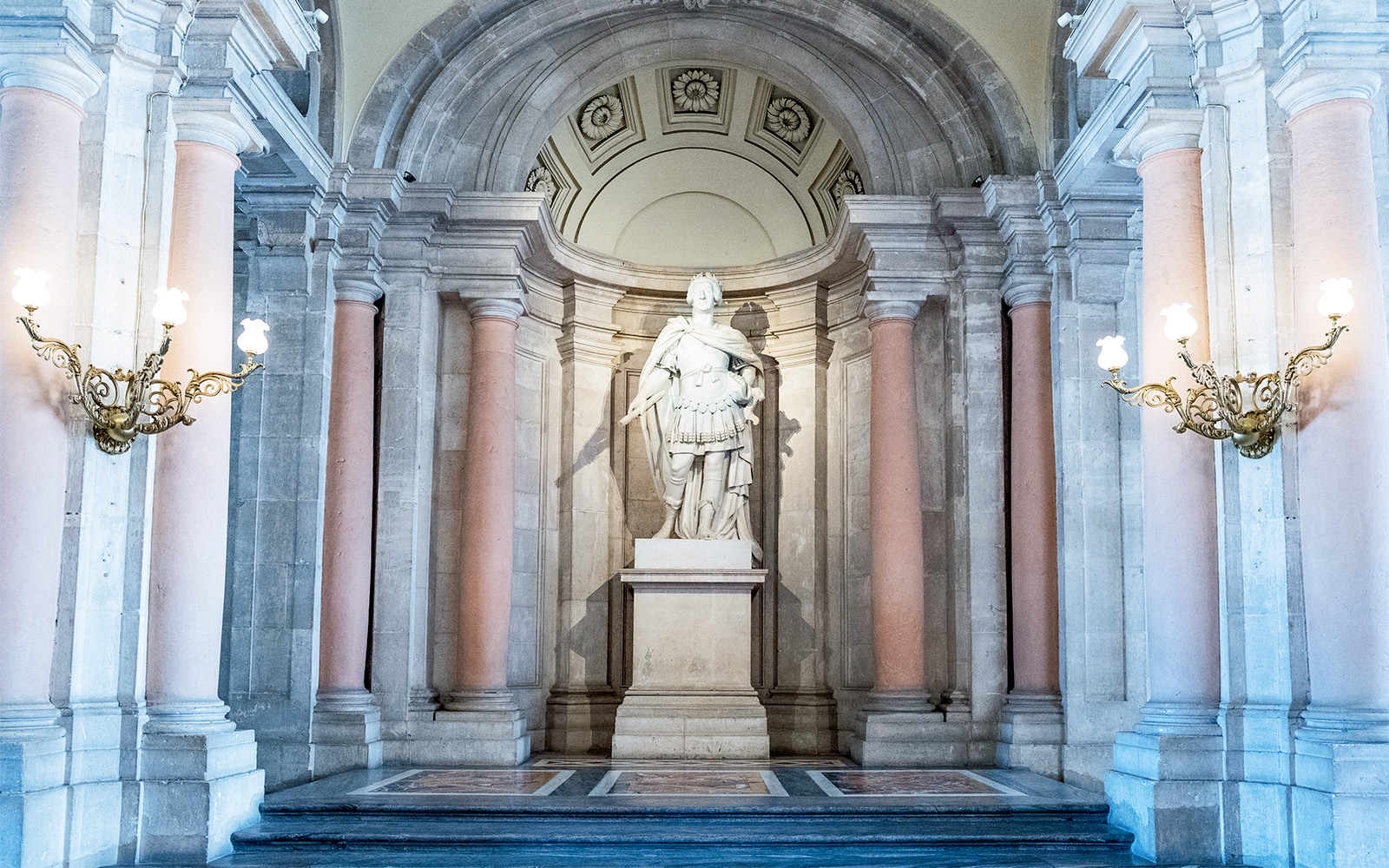 Statue in the Royal Palace of Madrid surrounded by ornate columns and lighting fixtures.