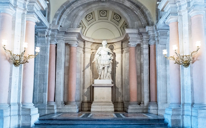 Statue in the Royal Palace of Madrid surrounded by ornate columns and lighting fixtures.