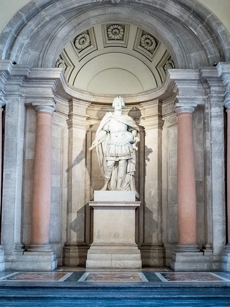 Statue in the Royal Palace of Madrid surrounded by ornate columns and lighting fixtures.