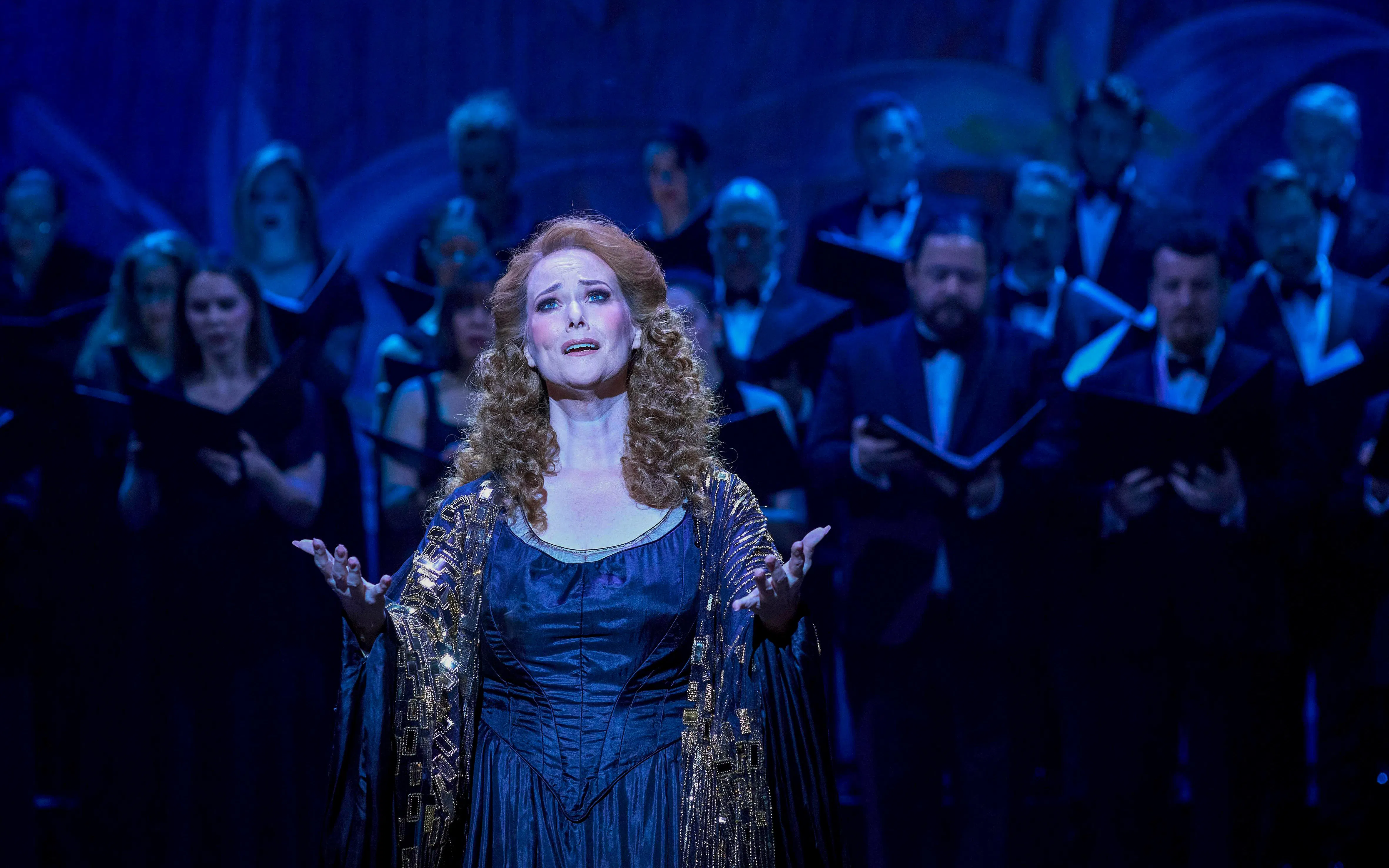 Performer singing on stage during a My Fair Lady production with a choir in the background.