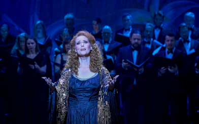 Performer singing on stage during a My Fair Lady production with a choir in the background.