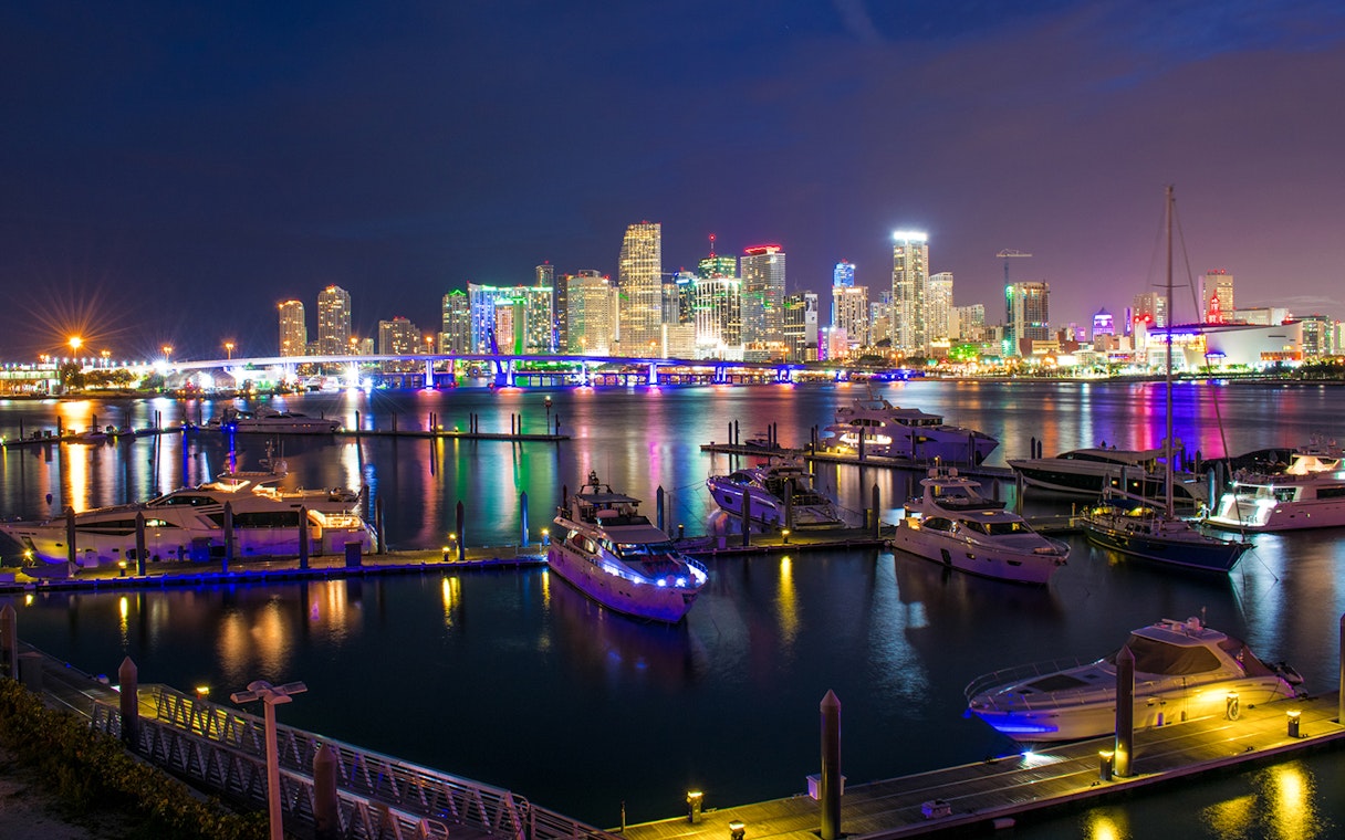 Miami skyline at night with illuminated buildings and yachts in the foreground.