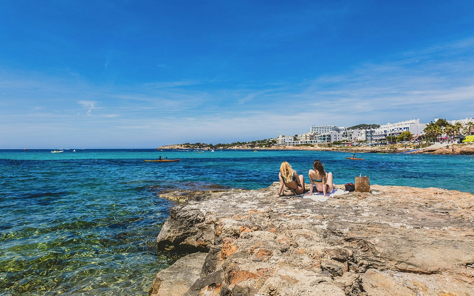 Two people relaxing on rocky shore overlooking turquoise waters at Sant Antoni de Portmany, Ibiza.