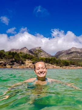 Person swimming in clear water at Wineglass Bay, Tasmania, with mountains in the background.