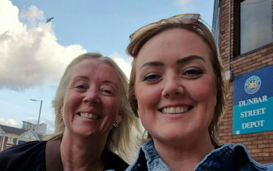 Two people smiling in front of Dunbar Street Depot sign, Belfast.