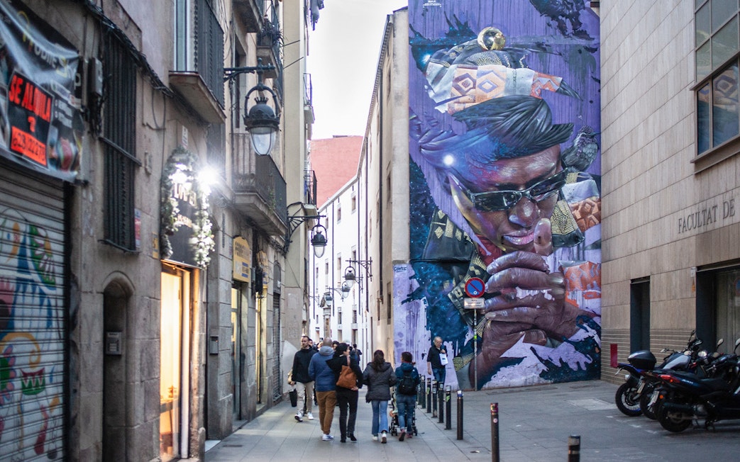 Street art mural in Raval, Barcelona, with people walking along a narrow alley.
