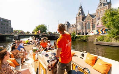 Guests enjoying drinks on a luxury open boat cruise in Amsterdam canal near historic buildings.