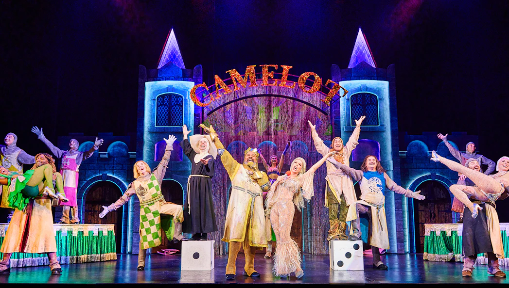 Performers on stage in a vibrant scene from Spamalot, with a castle backdrop and "Camelot" sign.