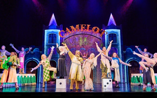 Performers on stage in a vibrant scene from Spamalot, with a castle backdrop and "Camelot" sign.