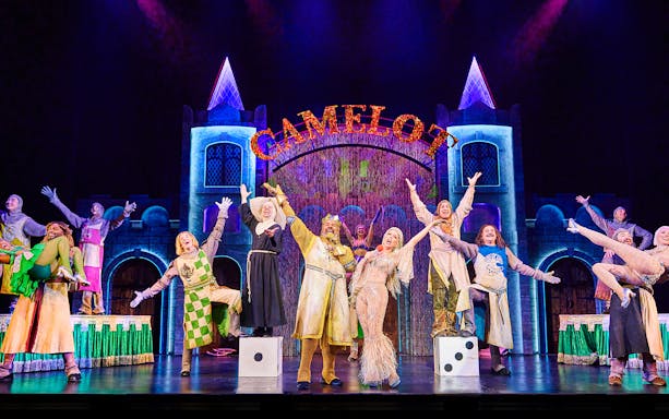Performers on stage in a vibrant scene from Spamalot, with a castle backdrop and "Camelot" sign.