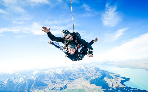 Skydivers tandem jumping over Mt Cook with snow-capped peaks and lake below.