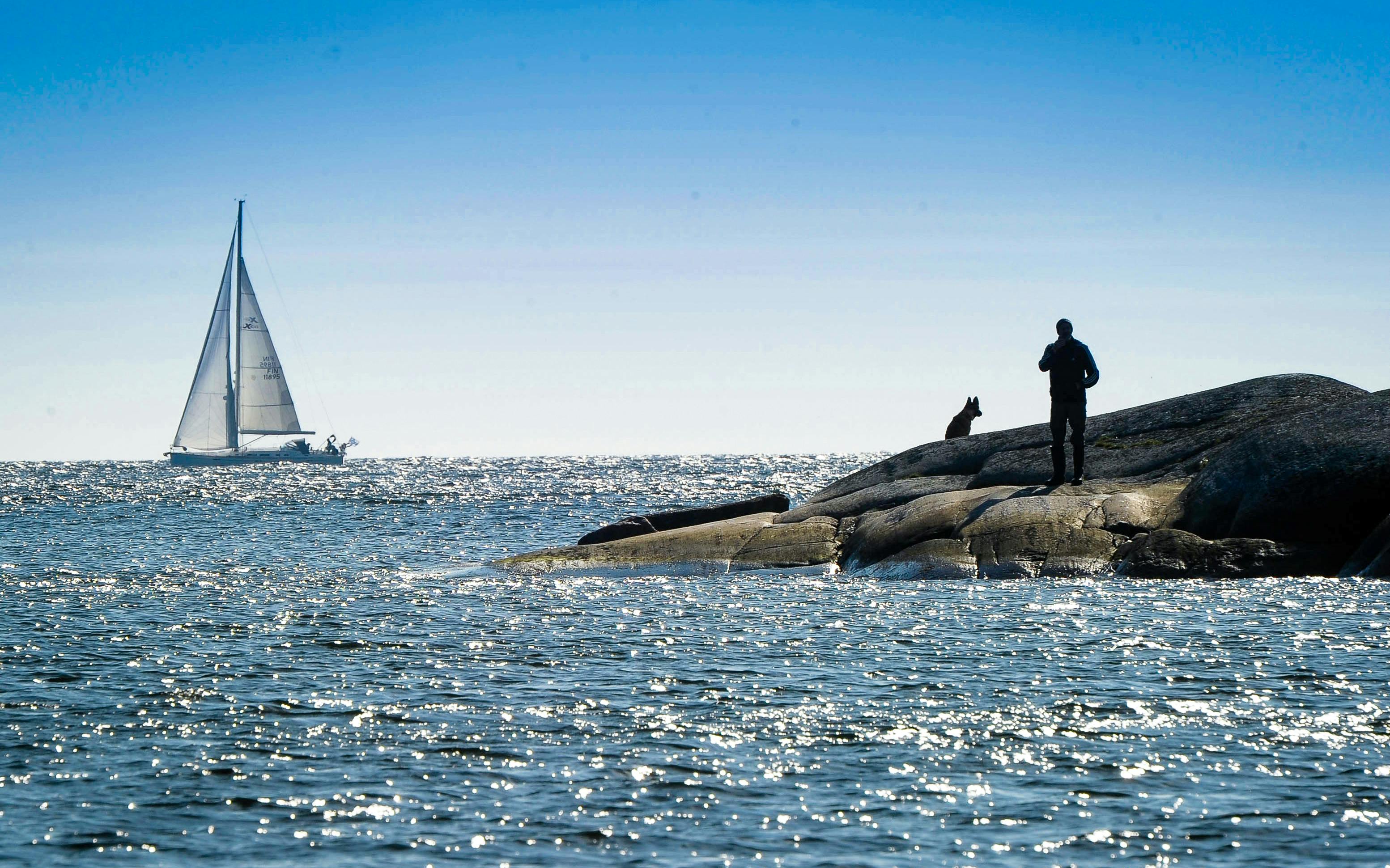 Sailboat on Helsinki archipelago waters with person and dog on rocky shore.