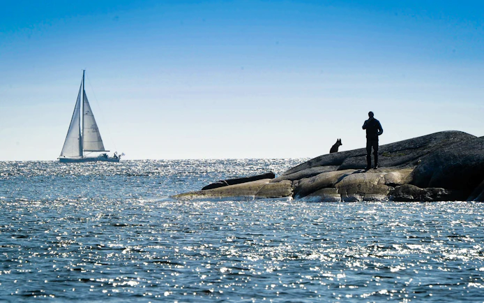 Sailboat on Helsinki archipelago waters with person and dog on rocky shore.