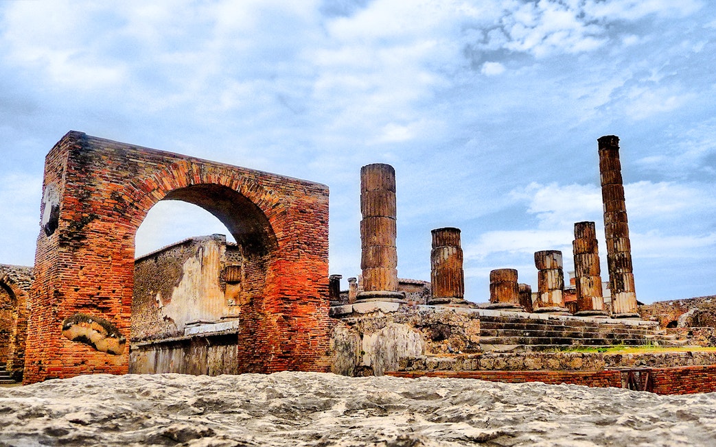 Pompeii ruins with ancient brick arch and stone columns under a blue sky.