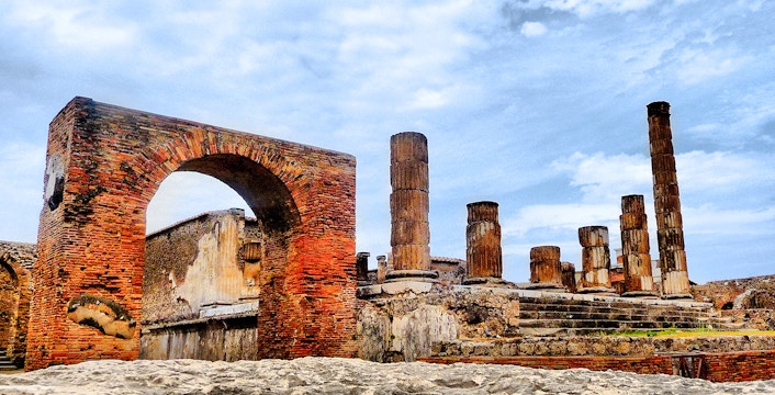 Pompeii ruins with ancient brick arch and stone columns under a blue sky.
