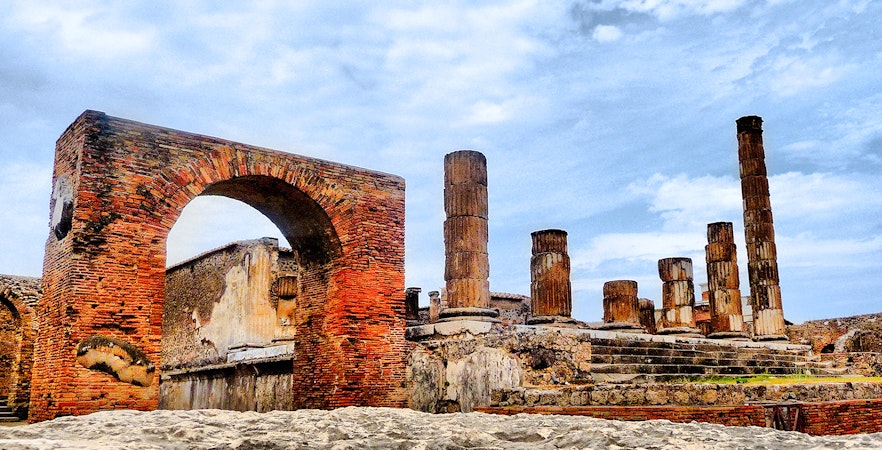 Pompeii ruins with ancient brick arch and stone columns under a blue sky.