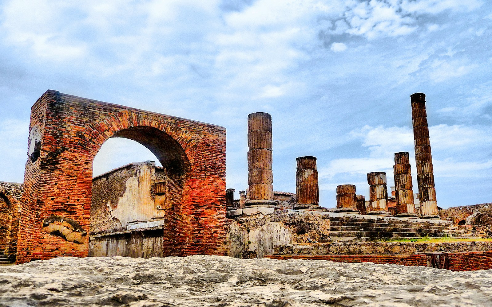Pompeii ruins with ancient brick arch and stone columns under a blue sky.