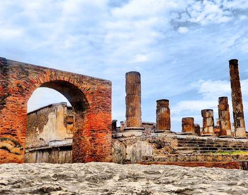 Pompeii ruins with ancient brick arch and stone columns under a blue sky.