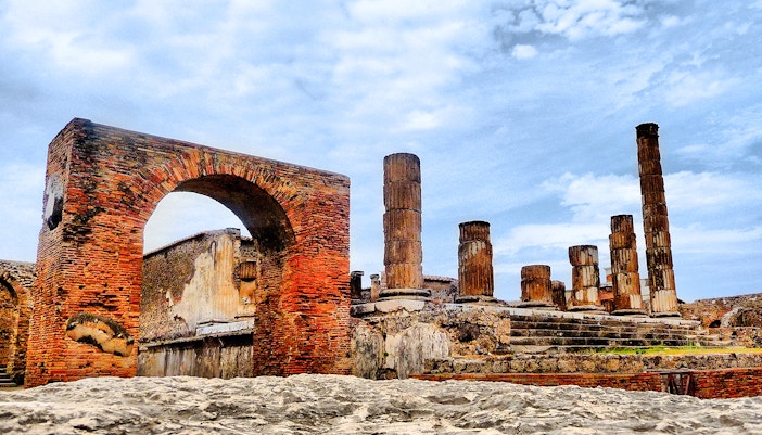 Pompeii ruins with ancient brick arch and stone columns under a blue sky.