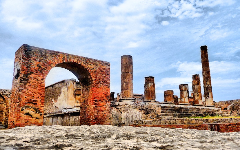 Pompeii ruins with ancient brick arch and stone columns under a blue sky.
