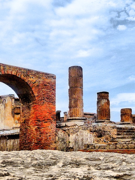 Pompeii ruins with ancient brick arch and stone columns under a blue sky.