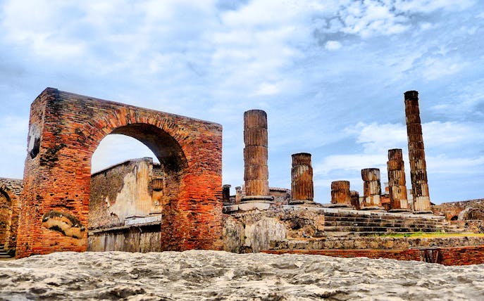 Pompeii ruins with ancient brick arch and stone columns under a blue sky.