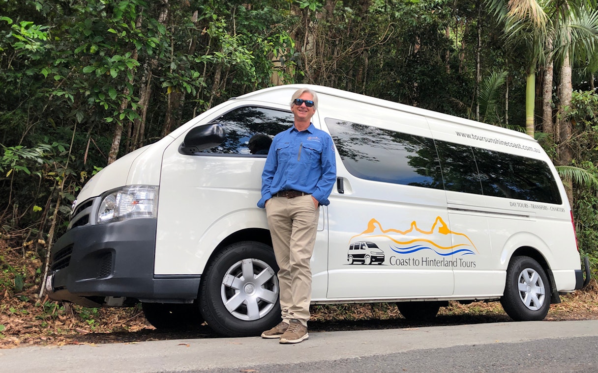 Tour guide standing beside a Coast to Hinterland Tours van for Eumundi Markets transfer.