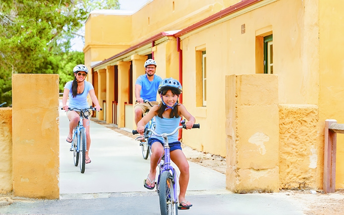 Family biking on Rottnest Island, passing historic yellow buildings.