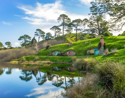 Hobbiton Movie Set with hobbit holes and lush green hills reflected in a pond, near Auckland.