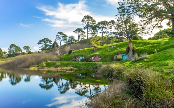 Hobbiton Movie Set with hobbit holes and lush green hills reflected in a pond, near Auckland.
