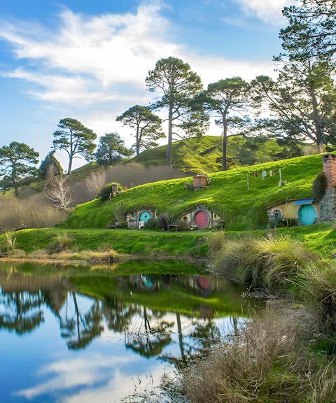 Hobbiton Movie Set with hobbit holes and lush green hills reflected in a pond, near Auckland.