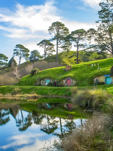 Hobbiton Movie Set with hobbit holes and lush green hills reflected in a pond, near Auckland.