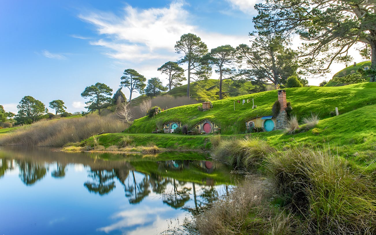 Hobbiton Movie Set with hobbit holes and lush green hills reflected in a pond, near Auckland.