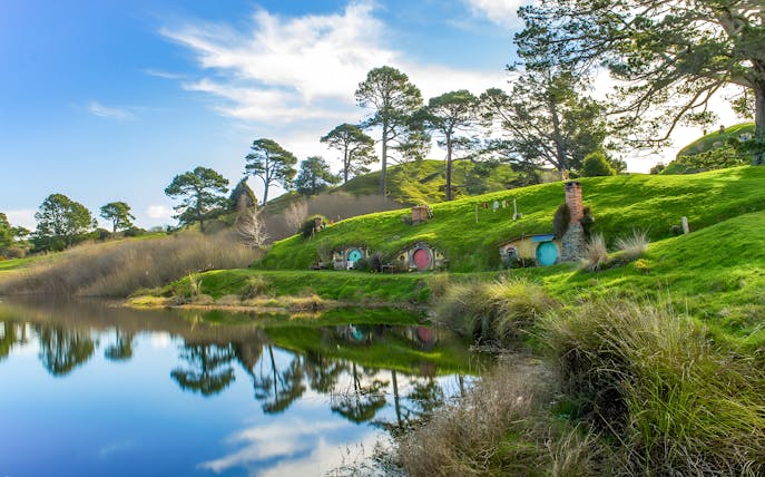 Hobbiton Movie Set with hobbit holes and lush green hills reflected in a pond, near Auckland.