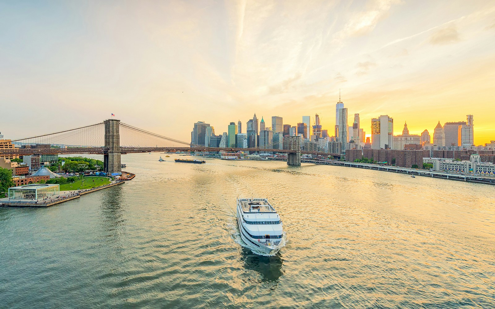 Cruise ship sailing at sunset with New York City skyline and Brooklyn Bridge in the background.
