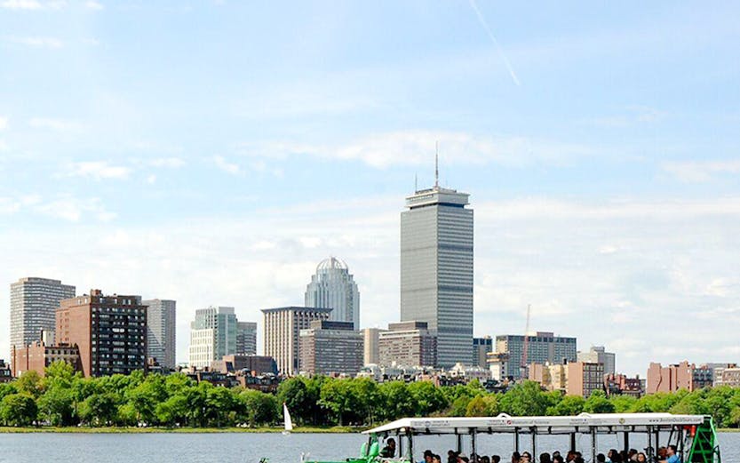 Green Boston Duck Tours boat cruising on Charles River with city skyline in background.