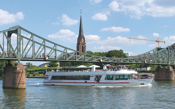 Cruise boat passing under a bridge in Frankfurt with a church spire in the background.