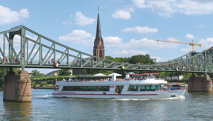 Cruise boat passing under a bridge in Frankfurt with a church spire in the background.