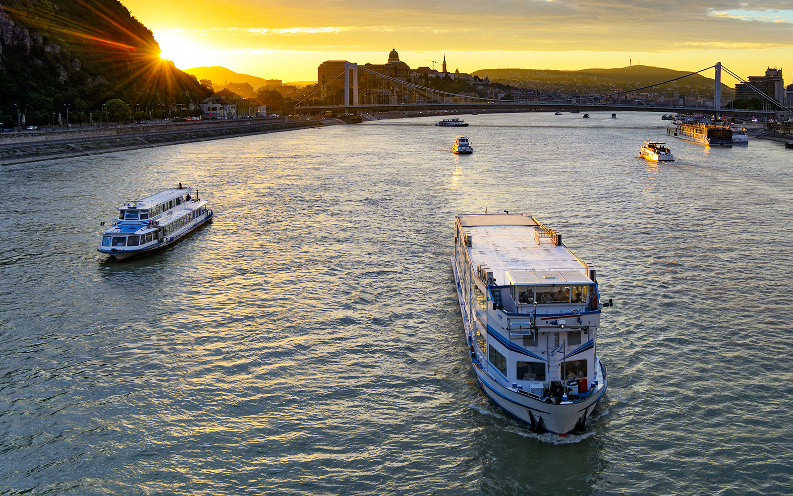 Cruise boat on Danube River at sunset with Budapest skyline in background.