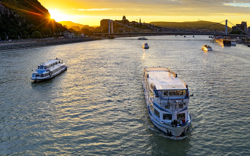 Cruise boat on Danube River at sunset with Budapest skyline in background.