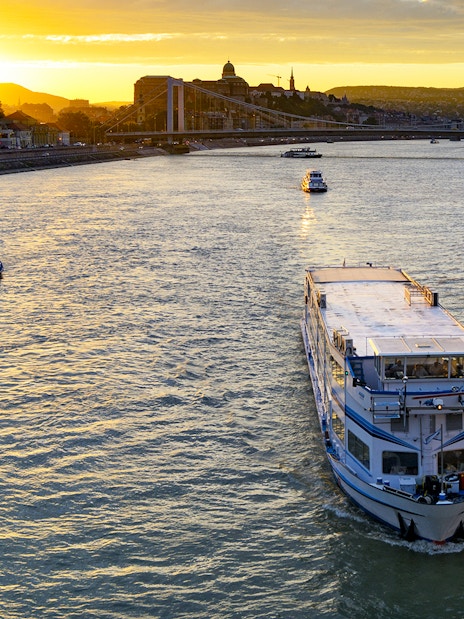 Cruise boat on Danube River at sunset with Budapest skyline in background.