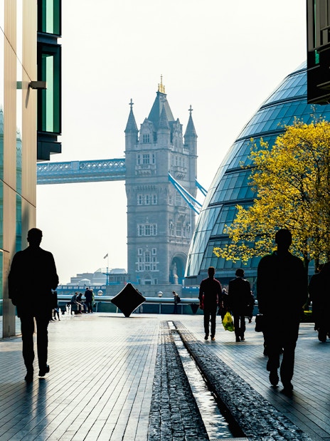 Silhouettes walking towards Tower Bridge, London, with modern buildings and trees.