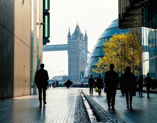 Silhouettes walking towards Tower Bridge, London, with modern buildings and trees.