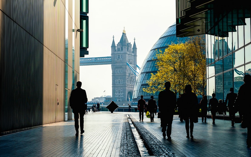 Silhouettes walking towards Tower Bridge, London, with modern buildings and trees.