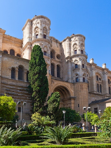 Málaga Cathedral facade with lush garden and tall trees in foreground.