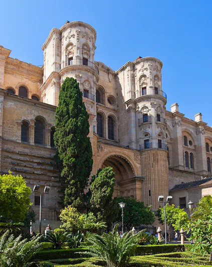 Málaga Cathedral facade with lush garden and tall trees in foreground.