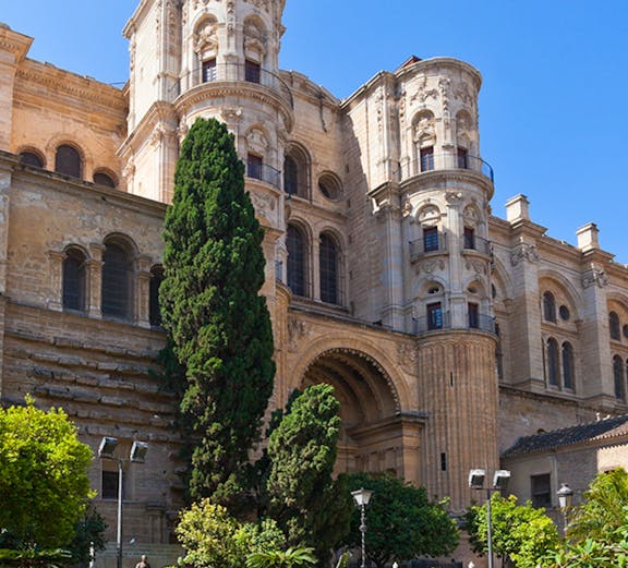 Málaga Cathedral facade with lush garden and tall trees in foreground.