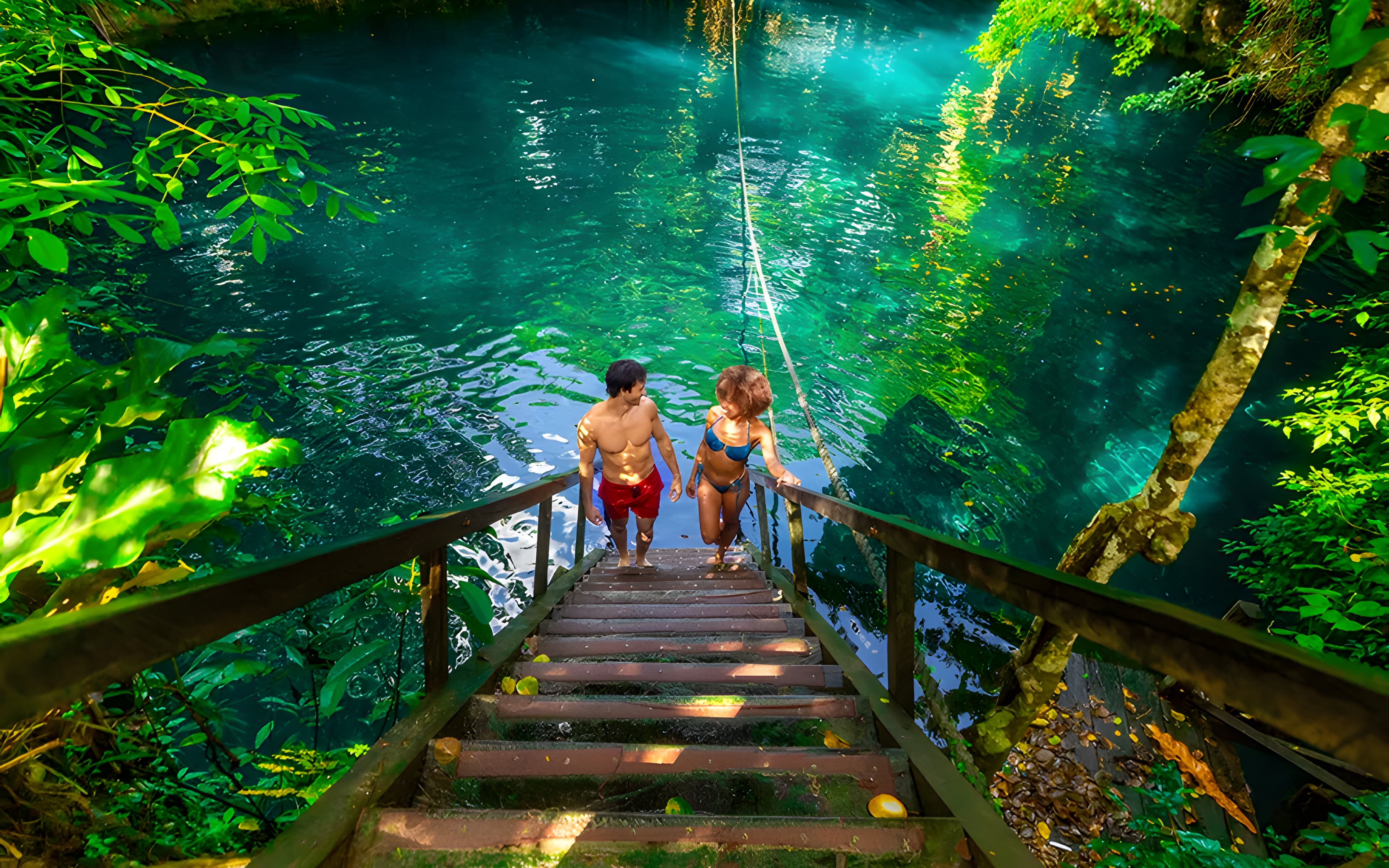 Couple ascending wooden stairs from a cenote surrounded by lush greenery.