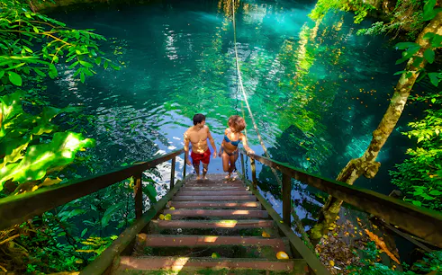 Couple ascending wooden stairs from a cenote surrounded by lush greenery.